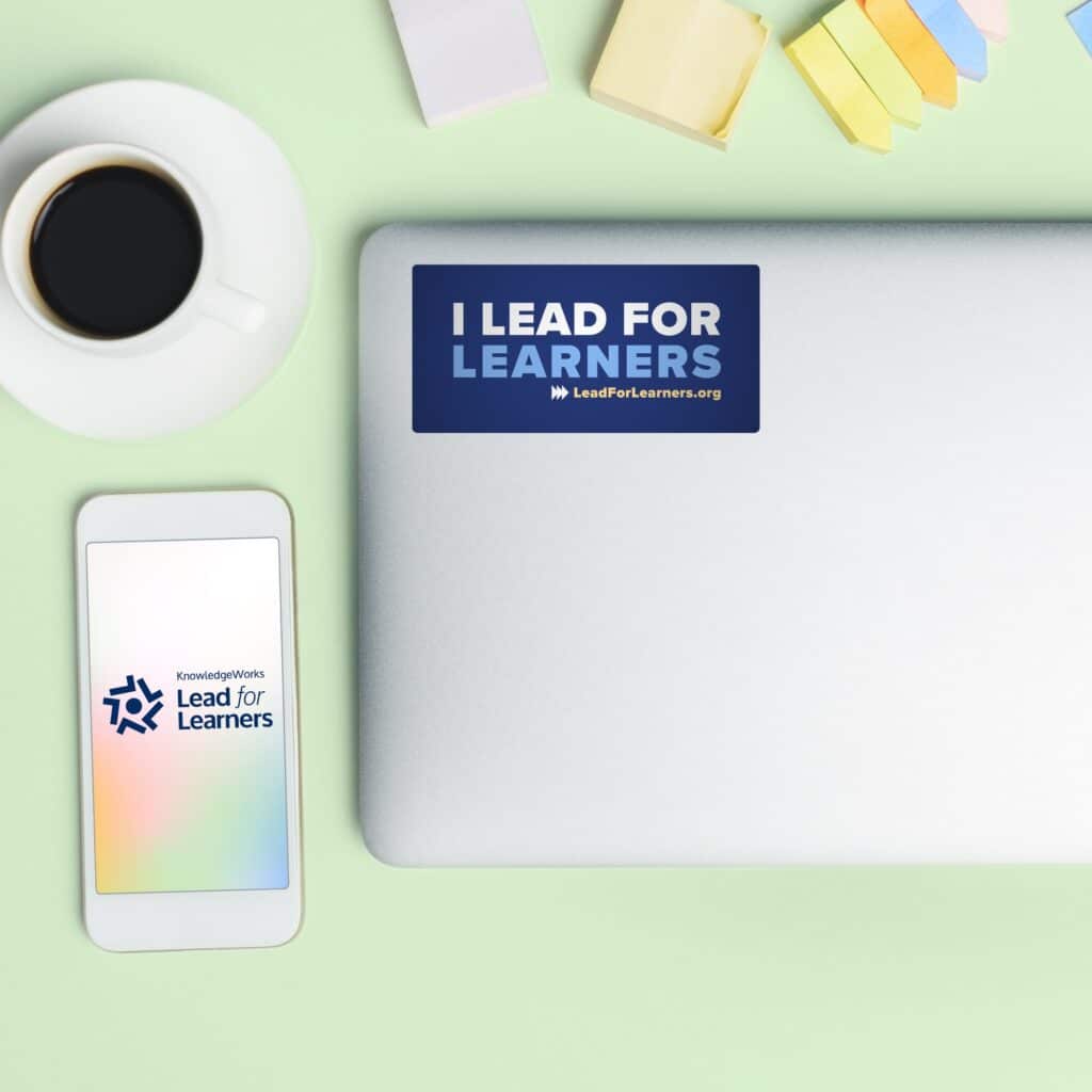 Laptop with "I Lead for Learners" sticker, phone with "Lead for Learners" app opening, a cup of coffee, and sticky notes on a green desk