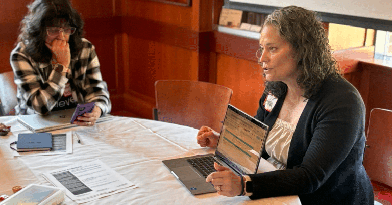 a woman with curly hair sits at a table with her laptop turned toward someone off-camera opposite her, showing her screen. A woman in the background is on the network app.