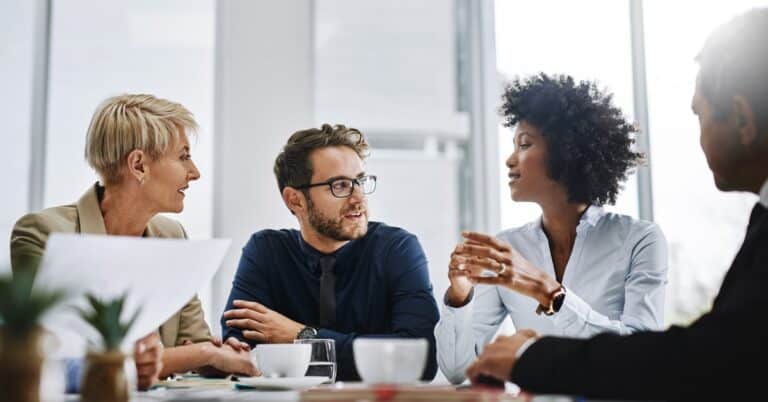 Adults of varying ages and skin color talk at a table papers and mugs in front of them