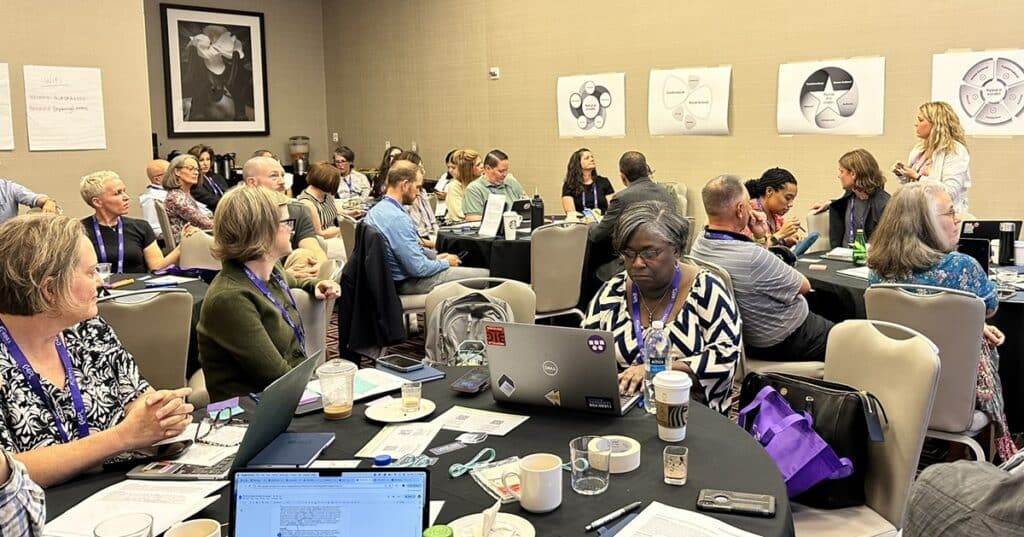 Educator gather in a large conference room across many tables. A woman standing facilitates.