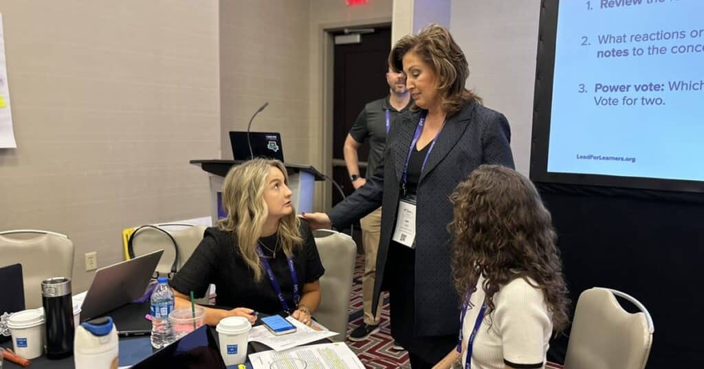 Two women sit at a table and speak to a woman standing behind them