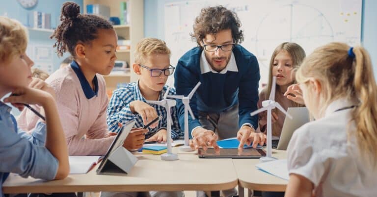 a male educator uses a tablet to show something to middle-school aged learners gathered around the table that has tiny windmills on it
