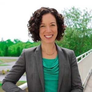 a portrait of Amy, a white woman with chin-length curly brown hair, wearing a necklace, sea green top and a gray blazer over it. She's leaning against a white railing of a bridge overlooking a stream of water lined by trees.
