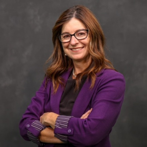 Portrait of Brenda, a white woman with wavy auburn hair down past her shoulders, wearing black rimmed glasses, pearl necklace and earrings, a black shirt and a purple blazer over it. She's smiling and standing in front of a gray background.
