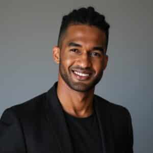Portrait of Donald, a young Black man with black hair, short on the sides and longer on top, pulled back in rows, and a five o'clock shadow. He's wearing a black shirt and black blazer over it, smiling at the camera in front of a gray background.