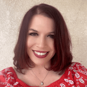 A portrait of Heather, a light-skinned woman with dark red, straight hair to her shoulders and brown eyes, smiling at the camera. She's wearing a necklace and a red patterned shirt and is standing in front of a cream colored wall.