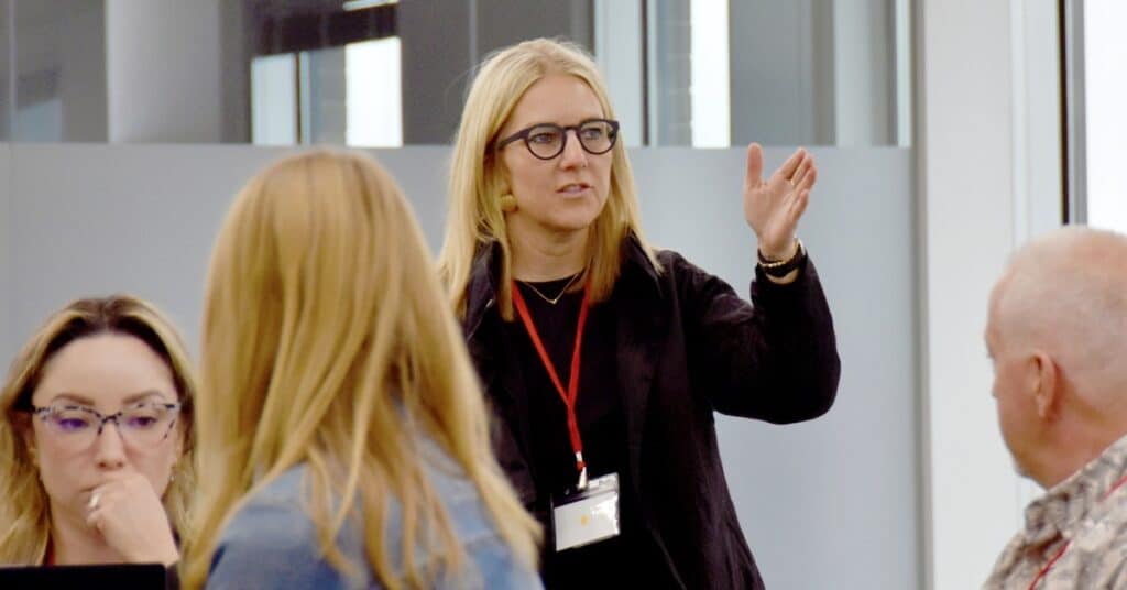 A blonde white woman stsands in front of a group sitting at tables with her hand out, animatedly talking