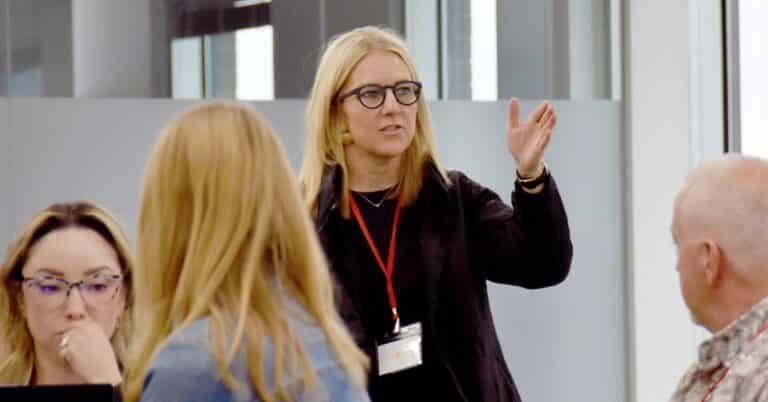 A blonde white woman stsands in front of a group sitting at tables with her hand out, animatedly talking