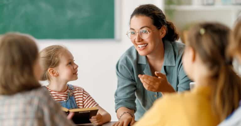 Woman in glasses leans over a table of children in a classroom to explain something with her hands