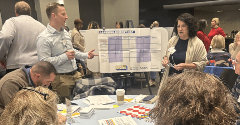 A group of adults gather in a large event space around a table, talking to each other in front of a chart on an easel